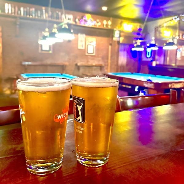 Two full beer glasses on a bar, with pool tables and neon lights in the background.